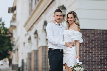 Fototapeta premium A smiling, stylish groom and a beautiful blonde bride in a white dress are hugging on the street in the city against the background of ancient buildings. Wedding photo of newlyweds in love.