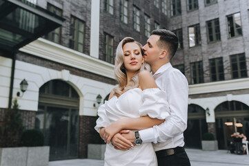 A smiling young groom and a beautiful blonde bride in a white dress are embracing on the street in the city against the background of modern, ancient buildings. Wedding photography of the newlyweds.