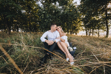 Naklejka na ściany i meble A young, smiling groom and a cute blonde bride with curly hair in a white dress are sitting on a rug on the yellow grass in the fall in nature and hugging. Wedding photo of beautiful newlyweds.