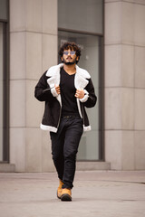 Curly indian man in sunglasses and jacket walking outdoors 