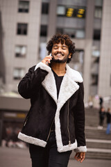 Fototapeta premium Happy indian man in jacket closing eyes while talking on smartphone in city 