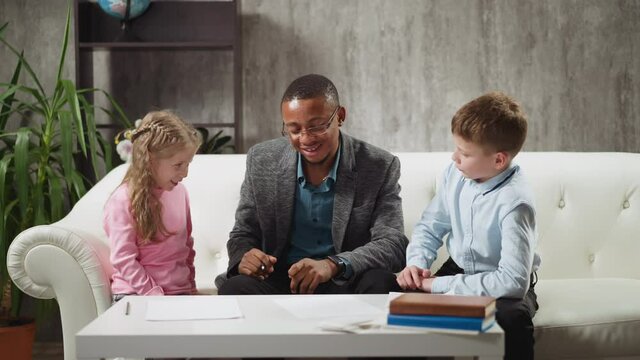 Young African-American man teaches kids to raise hand and listens answers during private English lesson with native speaker at home slow motion
