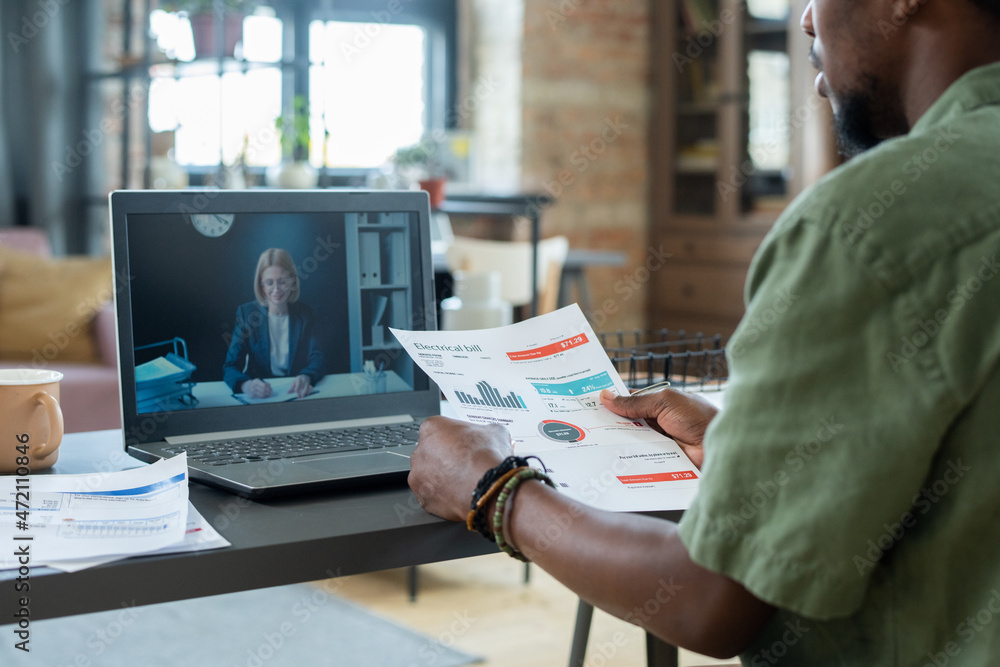Poster Over shoulder view of blackman in green shirt sitting at table and using laptop while discussing financial papers with advisor online