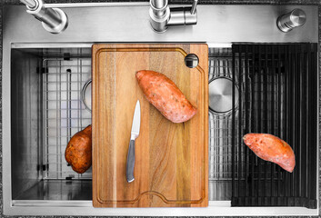 Three sweet potatoes ready to be sliced on wooden cutting board of workstation kitchen sink with...