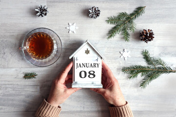 Calendar for January 8: hands hold a decorative house with the name of the month and the numbers 0 and 8, a cup with hot tea, snowflakes, fir branches on a gray background, top view
