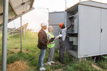 African american electrician in safety helmet and glasses checking voltage levels of solar panels in switchgear. Muslim woman and indian man using clipboard and laptop while standing behind.