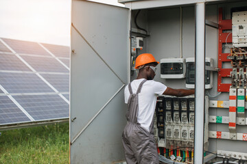 African american man in grey overalls and orange helmet repairing cables in switchgear at solar station. Concept of people, maintenance and alternative energy.