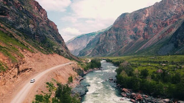 A white SUV jeep car is driving along an empty country road in the huge Katu-Yaryk canyon Chulyshman Valley with a large mountain bubbling river. Altai, Siberia, Russia