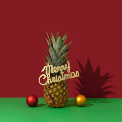 Pineapple decorated as christmas tree with merry christmas sign, red and golden baubles on vivid scarlet red and green background. Christmas celebration creative concept. Close up perspective.