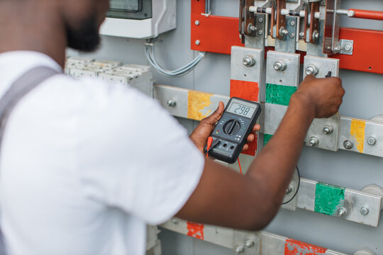 Close up of african american man standing near switchgear compartment with multimeter in hands. Competent technician controlling working process of solar station.