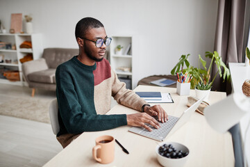 Portrait of young African-American man wearing glasses while working from home and using laptop,...