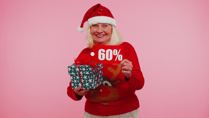 Grandmother woman in Christmas sweater showing gift box and 60 Percent discount inscriptions banner text. Black Friday. Good holiday low price for shopping. Blue studio wall background. Happy New Year