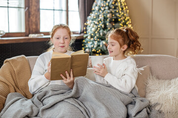 Two sisters are sitting on the couch and reading Christmas stories. Delicious hot cocoa.