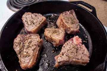 Close up view of several juicy lamb chops sizzling in a hot cast iron skillet on a white stove top