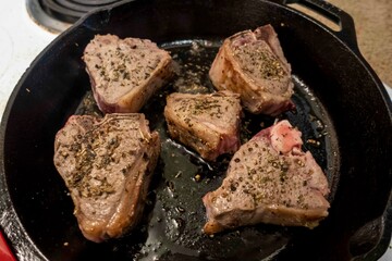Close up view of several juicy lamb chops sizzling in a hot cast iron skillet on a white stove top