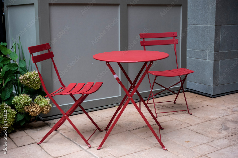 Wall mural isolated view of two red chairs at a red table outdoors on a cement brick patio, with lush green fol