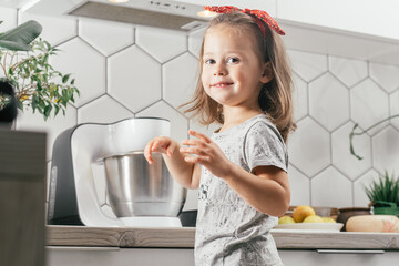 Little dark-haired girl 3 years old in headband bakes apple pie in kitchen. Child stands near planetary mixer and smiles