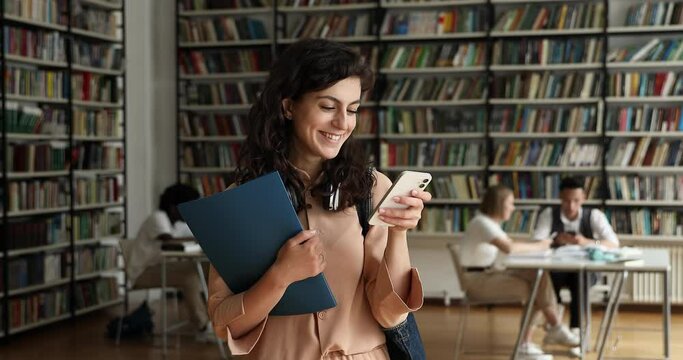 Attractive student girl standing in library holds cell phone, texting message, chatting in social media, check on-line studies timetable using mobile application, gen Z and modern tech usage concept
