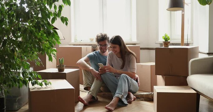 Family couple sit on floor at new rented purchased home choose future interior design online via phone. Happy young spouses relax among boxes with belongings at moving day plan renovation using cell