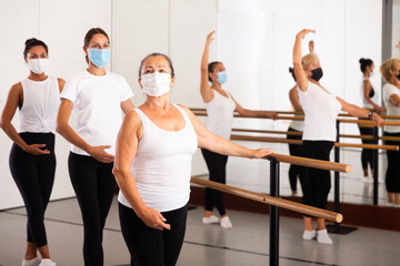 Dancing women in protective masks, engaged in ballet in the studio during the pandemic, stand holding the barre i