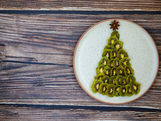 Funny edible dry kiwi chips creative christmas tree on ceramic plate - healthy christmas or new year snack idea for kids party, top view flat lay, close-up, on wooden background