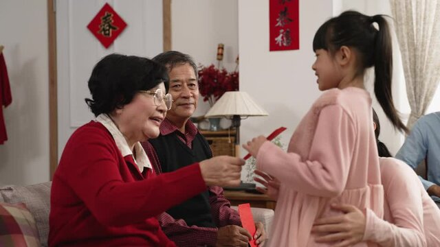 smiling asian grandfather and grandmother gesturing at granddaughter to come and giving lucky money to her in the living room on chinese new year. text translation: “spring” and “congratulations”