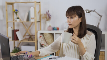 Obraz na płótnie Canvas gorgeous asian korean female entrepreneur with cup of coffee in hand is replying an important email on notebook