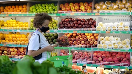 Curly woman in a face mask is choosing fruits in supermarket during coronavirus shopping 
