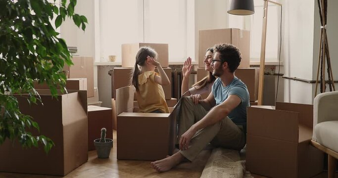 Happy young family couple becoming homeowners after buying new flat take break in unboxing packs with belongings at moving day sit on floor play patty cake with cute little daughter getting out of box
