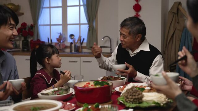 smiling asian senior man grandfather talking with hand gestures as the girl granddaughter in red dress giving food to him at the family reunion dinner party on the eve of lunar new year