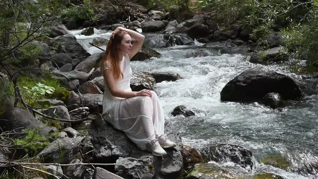 Portrait of long-haired beautiful woman in white dress relaxing on mountain river bank in summer forest. Romantic young girl sits alone on big stone, enjoys beauty of wild nature.