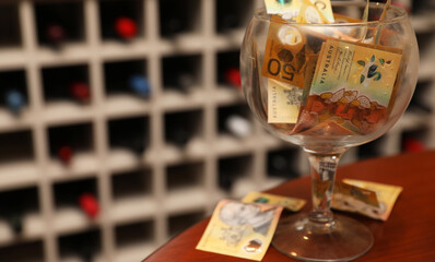 A large clear wine glass filled with fifty dollar notes on a timber table with a wine cellar or cabinet filled with bottles in the background.