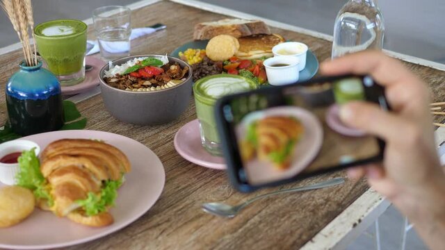 Hand of trendy blogger taking a picture of healthy breakfast with croissant sandwich, smoothie bowl, toasts, salad and matcha latte