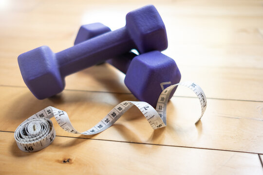 A White Measuring Tape Laying Next To Two Purple Dumbbells On The Wooden Floor Of A Home Gym