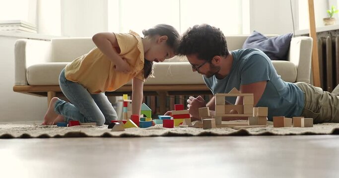 Caring single dad lie on carpet at playroom enjoy building city from bricks with small daughter. Grownup elder brother play on warm floor with sister preschooler help create toy town from wooden parts