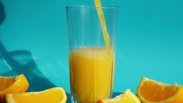 Pouring orange juice stream from jug into glass of squeezing Orange juice with sliced fruits on blue background. Fruit juice, fresh cold drink. Refreshment in summer. Fruits cut in half. Copy space
