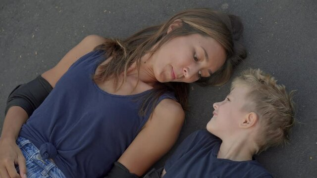 From-above medium close-up of happy young Caucasian woman and her cute 7-year-old son wearing elbow pads lying on ground in skatepark having fun together