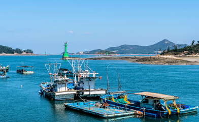 Fishing trawlers anchored in small port with green lighthouses in background.
