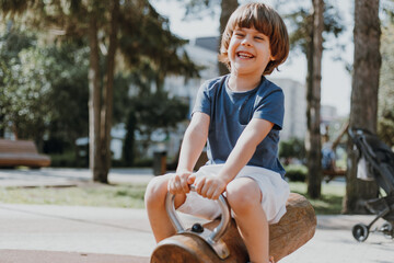 happy laughing little boy in a blue T-shirt and white shorts rides on a wooden rocking chair outdoor. child is playing on a public playground on a sunny summer day. lifestyle. space for text