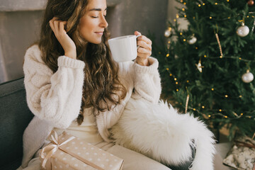Young woman drinking hot coffee or cocoa sitting on couch in cozy room with Christmas tree. Winter...