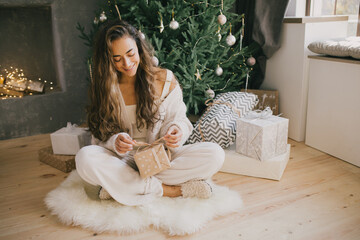 Young beautiful woman holding gift box in a cozy room with Christmas tree and presents on background.