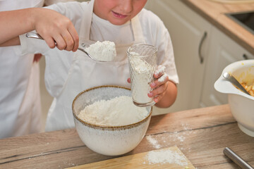 A boy in chef clothes pours flour into the dough for cooking apple pie