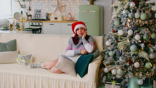 Full Body Of Annoyed Female In Santa Hat And Warm Wear Sitting On Sofa With Bad Wrapped Present Near Christmas Tree In Studio Apartment Decorated With Glowing Garlands