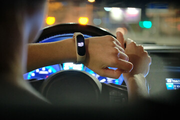 Close up of driver hand with digital watch checking time holding steering wheel driving car with blurred city street lights on background at night