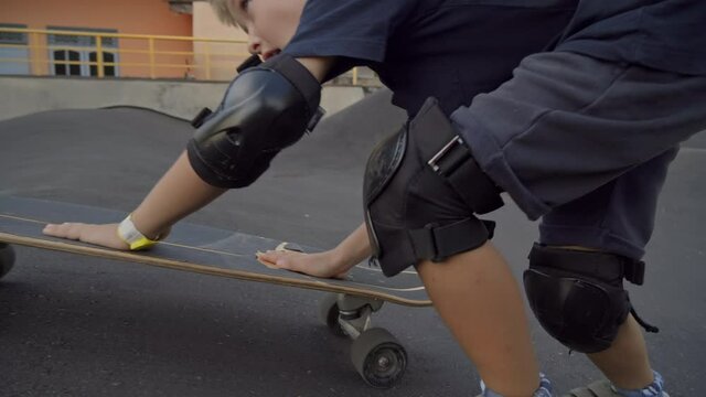 Tracking shot of active 7-year-old boy in knee and elbow pads pushing and running after skateboard rolling down skateboarding rink outdoors