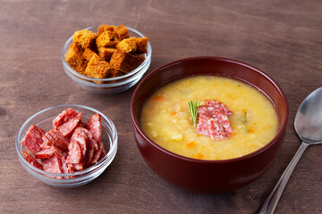 vegetable soup with meat and crackers. Split pea soup with ham, carrots and potatoes on a rustic wooden background. Classic Dutch pea soup erwtensoep, snert with bacon in the brown bowl on the table.