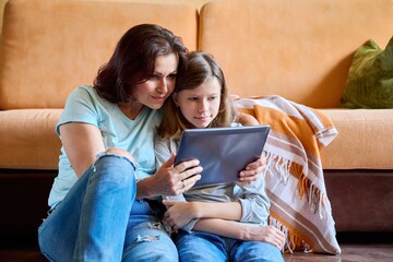 Middle-aged mom and preteen daughter relaxing at home together, with digital tablet