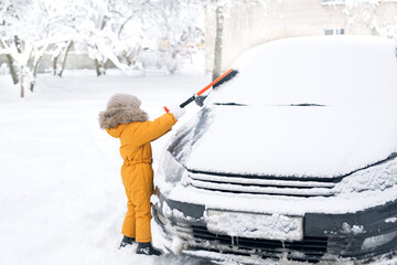 Child sweeps snow from the windshield of a car with a brush after a heavy snowfall. Kid cleans the...