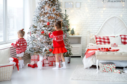 Kids hang Christmas toys and balls on an artificial Christmas tree in a white bedroom. Gifts in holiday packaging lie under a spruce tree. Girls are dressed in red sweaters. Decorating a tree for xmas