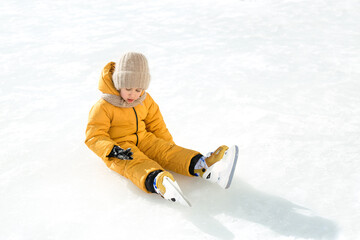 Fototapeta na wymiar The girl fell on the ice, taking her first steps on skates. A child learns to skate. A child sits on an ice rink and examines mittens that were injured during the fall. Ice skating season on the pond 